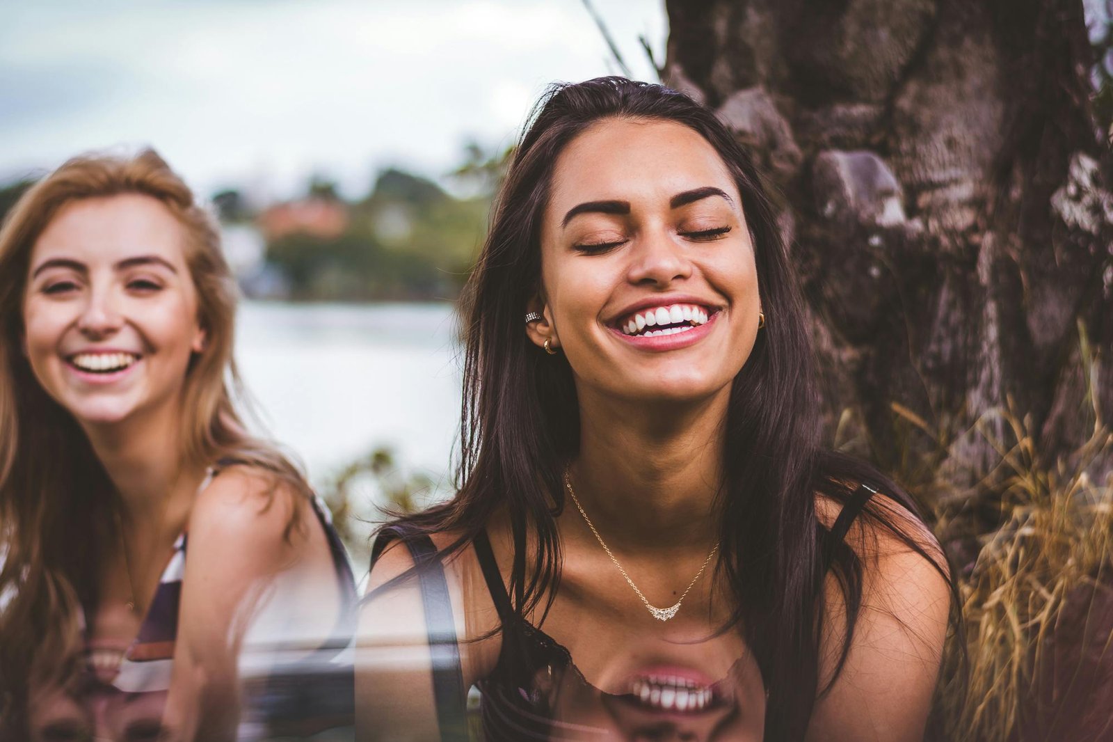 Two women happily laughing together outdoors by a serene lakeside.